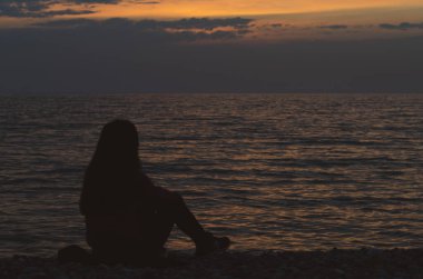 Gün batımında sahilde oturan bir kızın silueti.