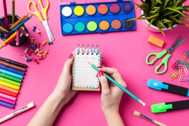 Kadın eli boş not defterinde yazılı. Pembe bir arka plan üzerinde okul ya da sanat malzemeleri