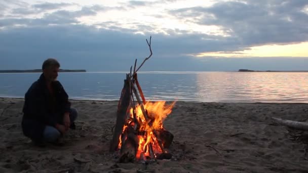 Hombre sentado junto al fuego cerca del río al atardecer — Vídeos de Stock