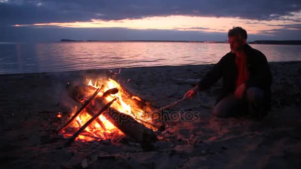 El hombre está lanzando leña a un incendio en la orilla del río al atardecer, Ob River, Siberia, Rusia — Vídeo de stock