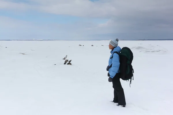 Vrouw met een rugzak in een blauwe jas staande op de oever van een besneeuwde rivier — Stockfoto