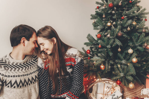 stylish hipster couple in sweaters hugging and embracing at Christmas tree in cozy evening room.