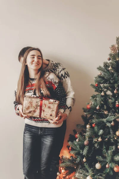 kucaklayan ve hediye kutusu ile Noel ağacı, kırmızı yay ayakta tutan kazak genç mutlu çift