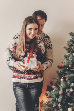 kucaklayan ve hediye kutusu ile Noel ağacı, kırmızı yay ayakta tutan kazak genç mutlu çift