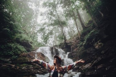 happy stylish traveler girl in hat relaxing at waterfall, atmospheric moment of success. hipster woman with backpack travelling, hands up. space for text. wanderlust and travel concept clipart