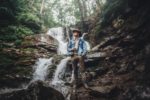 癖和旅游理念 时尚的旅行者在瀑布 手持照相相机 探索树林的帽子姿势 背包旅行的时髦人在森林里 — 图库照片