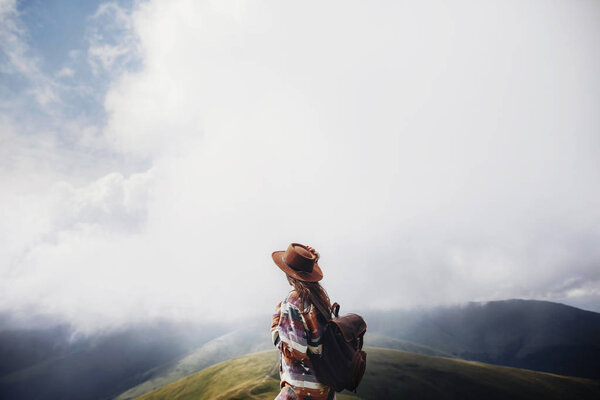 wanderlust and travel concept. girl traveler in hat with backpack looking at clouds in mountains. stylish hipster woman exploring on top of mountain. space for text. atmospheric moment