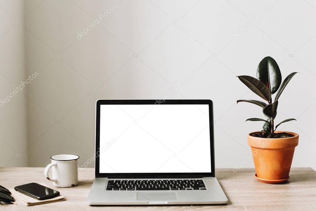 Laptop with white blank screen on wooden desktop with phone, notebook, coffee and plant. 