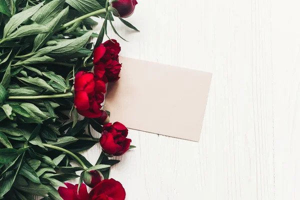 白い木製の素朴な背景 フラットの美しい赤牡丹花束とクラフト紙のカードを置きます テキストのためのスペースとモダンなグリーティング カード トップ ビュー — ストック写真