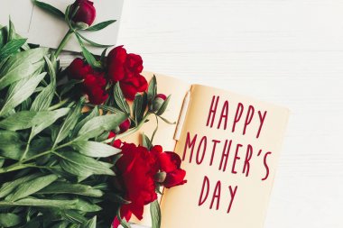 mutlu anneler günü metni imzalamak craft kağıt kartında Beyaz ahşap rustik arka planda, düz güzel kırmızı peonies yatıyordu. Modern tebrik kartı. Anneler günü. şık çiçek açan çiçekler