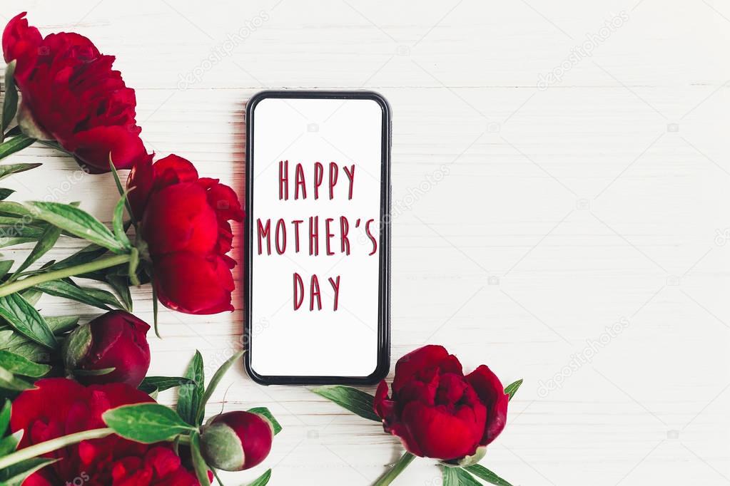 happy mother's day text sign on screen phone and beautiful red peonies on white wooden rustic background, flat lay. modern greeting card. mothers day. stylish blooming flowers