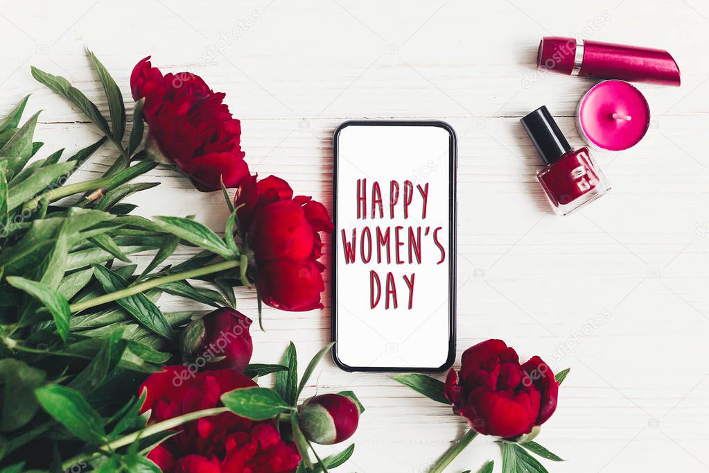 happy women's day text sign on smart phone empty screen and beautiful red peonies on white wooden rustic background, flat lay. modern greeting card. womens day