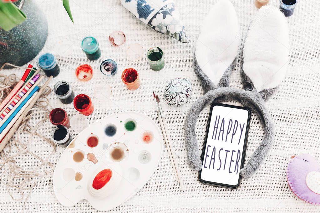 Happy Easter text on phone screen. season's greetings card. bunny ears and smartphone and colorful egg on rustic background flat lay. happy easter concept