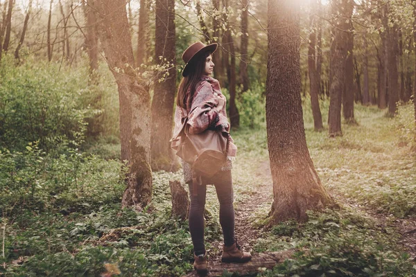 时尚时髦的旅行女孩在帽子与背包漫步在森林中惊人的傍晚阳光灯 — 图库照片