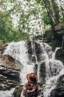 şelale orman dağlarında bakarak sırt çantası ile şapkalı şık hippi kız. Gezgin kadın ormanda keşfetmek. seyahat ve Yolculuk tutkusu kavramı. Atmosferik an