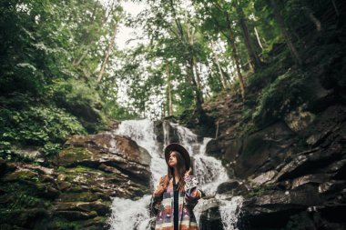şelale orman Dağları'nda, sırt çantası ayakta ile şapkalı şık hippi kız. Gezgin kadın ormanda keşfetmek. seyahat ve Yolculuk tutkusu kavramı. Atmosferik an