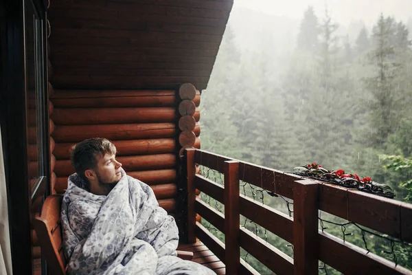 stylish bearded man relaxing on wooden porch among forest in rainy mountains. hipster guy resting in blanket, sitting in cottage, looking at rain. calm peaceful moment
