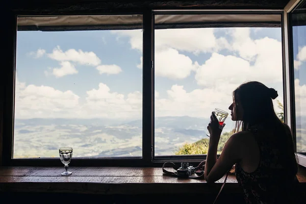 时尚的女孩旅行者在窗口在小屋 在山上和天空的看法饮用 快乐的时髦女人拿着玻璃和放松 暑假的概念 旅行和流浪 — 图库照片