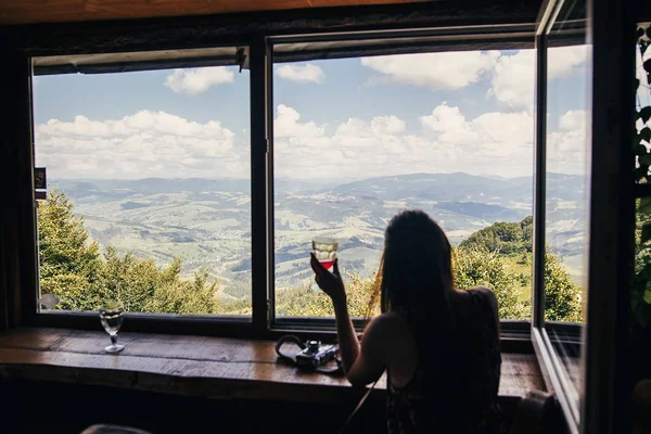 时尚的女孩旅行者坐在窗口与风景在山和天空 快乐的时髦女人拿着饮料 看着天空 暑假的概念 关注高山 — 图库照片