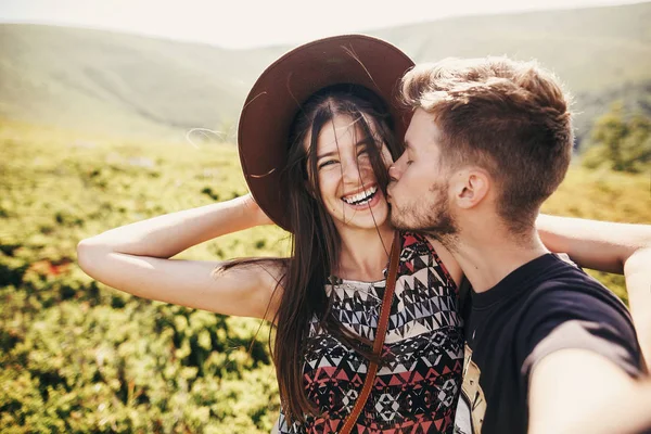 スタイリッシュな流行に敏感なカップル Selfie をとり 日当たりの良い夏の日に山の上に笑みを浮かべてします 美しい男とハグし 山帽子で幸せな女 一緒に旅行します — ストック写真