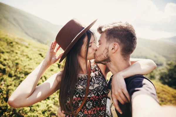 スタイリッシュな流行に敏感なカップル Selfie をとり 日当たりの良い夏の日に山の上にキスします 美しい男と帽子を受け入れ幸せな女が一緒に旅行します — ストック写真