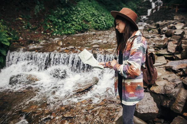 时尚时髦的女孩在帽子与背包探索地图 站在河与瀑布在山上 旅行的女人在树林里旅行 旅行和流浪概念 大气力矩 — 图库照片