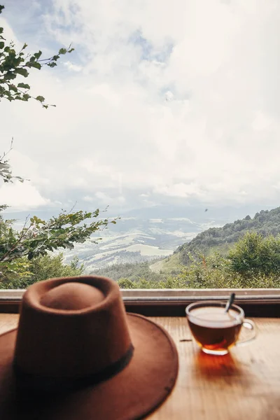 时髦的帽子和茶在玻璃上的木桌上 在窗口的光与山和天空的看法 山顶上有美味的热饮料 夏天旅行和流浪 关注高山 — 图库照片