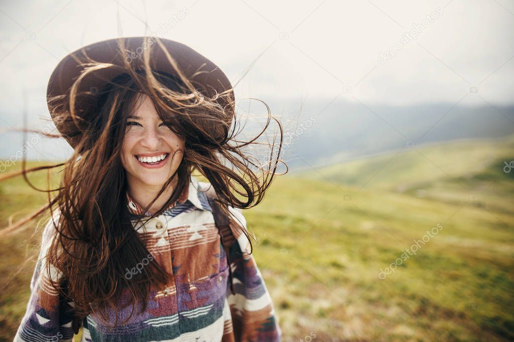 happy traveler hipster girl with windy hair  smiling, standing on top of sunny mountains. space for text. stylish woman in hat. atmospheric moment. travel and wanderlust. space for text