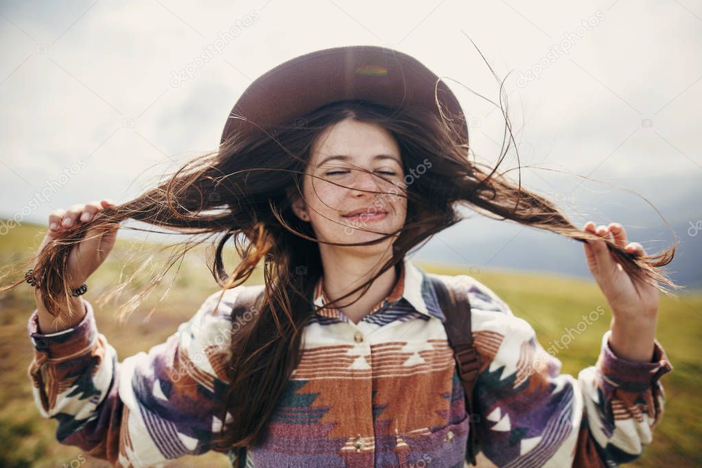 happy traveler hipster girl with windy hair  smiling, standing on top of sunny mountains. space for text. stylish woman in hat holding hair. atmospheric moment. travel and wanderlust