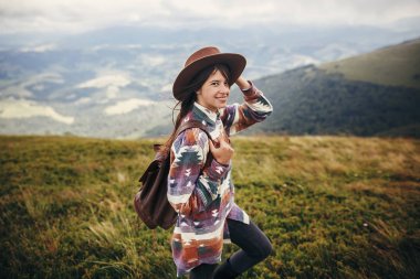 happy stylish traveler girl holding hat with backpack and windy hair in mountains clouds. summer vacation. travel and wanderlust concept. space for text. amazing atmospheric moment clipart