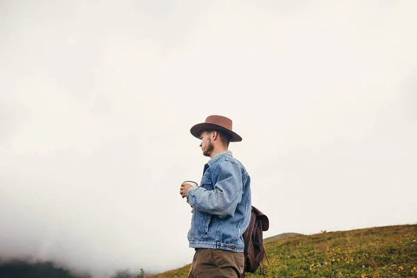 旅行和流浪概念 时尚的旅行者在帽子与背包捧着杯茶在山上云 文本的空间 时髦的家伙旅行 惊人的大气时刻 — 图库照片
