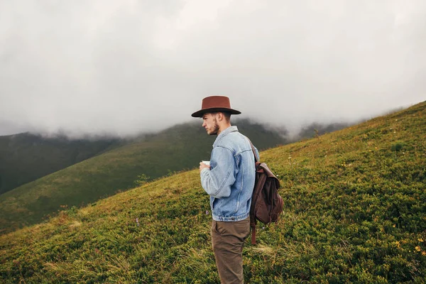 时尚的旅行者在帽子与背包捧着杯茶在山上云 文本的空间 时髦的家伙旅行 惊人的大气时刻 旅行和流浪概念 — 图库照片