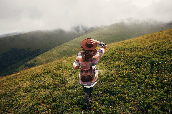 stylish traveler girl in hat with backpack looking at sunny mountains in clouds. summer vacation. travel and wanderlust concept. space for text. back view. amazing atmospheric moment