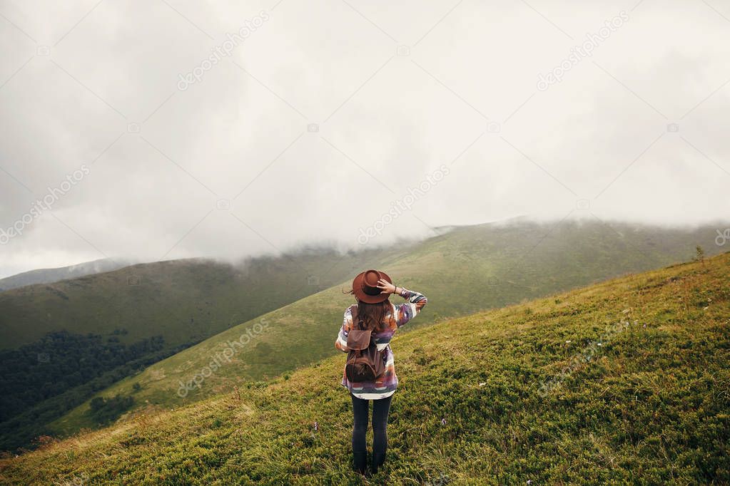 stylish traveler girl in hat with backpack looking at sunny mountains in clouds. summer vacation. travel and wanderlust concept. space for text. back view. amazing atmospheric moment