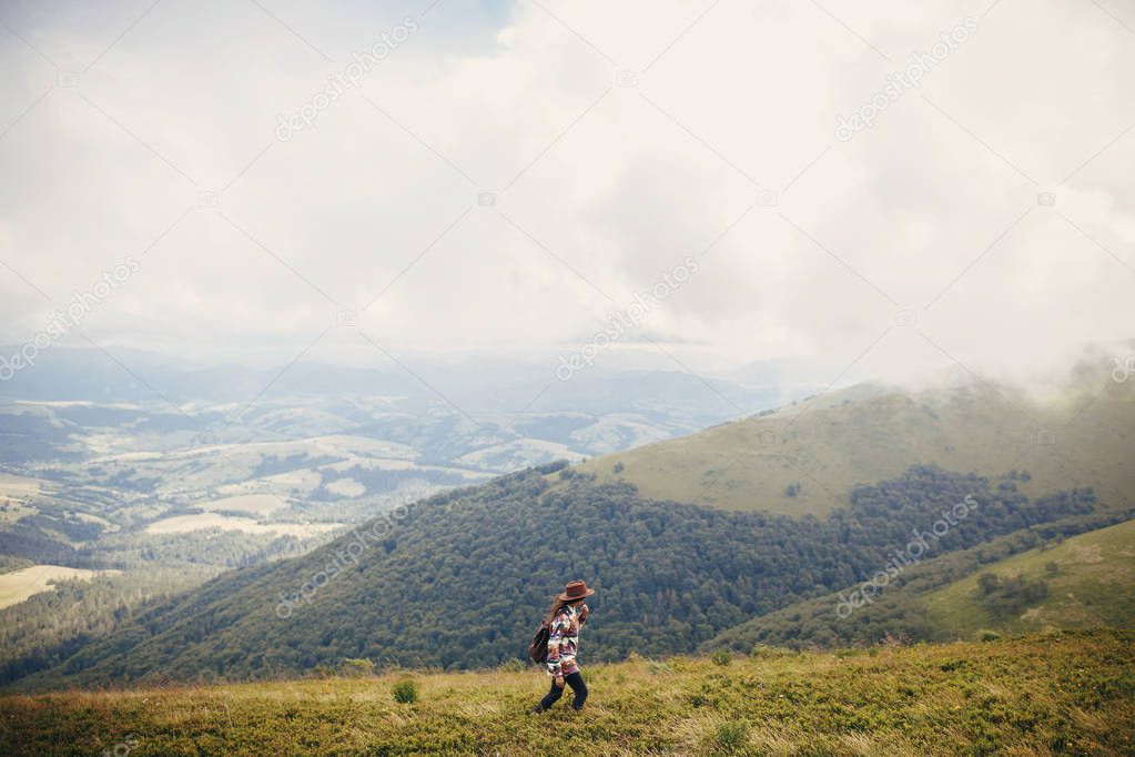 happy stylish traveler girl holding hat with backpack and windy hair in mountains clouds. summer vacation. travel and wanderlust concept. space for text. amazing atmospheric moment