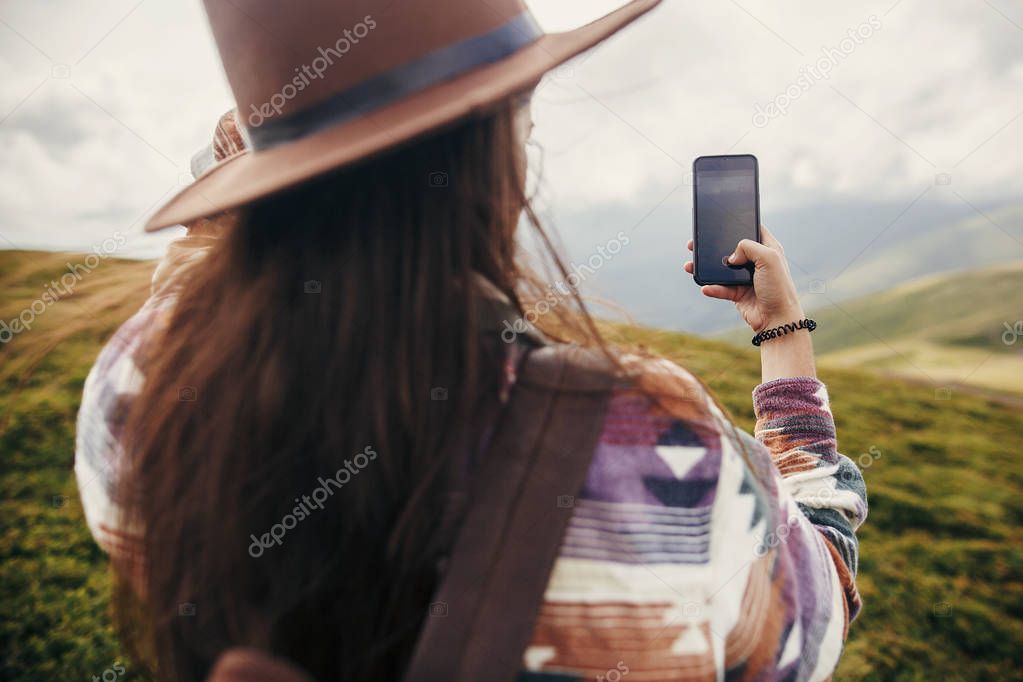 stylish traveler girl in hat with windy hair holding phone and taking picture on top of  mountains. 