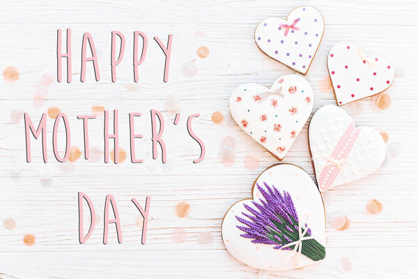happy mothers day text with pink cookies hearts and purple flowers on white rustic wooden background with confetti in soft light.