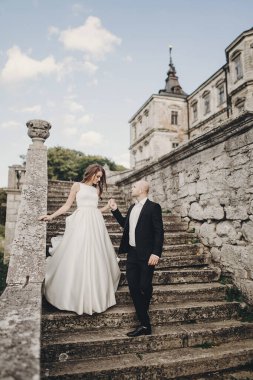 Muhteşem bir düğün çifti parktaki eski şatonun yakınındaki taş merdivenlerde yürüyor. Eski binanın arka planında poz veren muhteşem gelinlik ve damatla şık bir gelin.