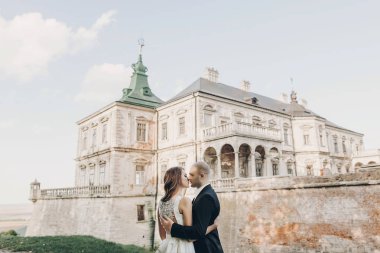 Güzel parktaki eski şatonun yakınında güneş ışığında kucaklaşan muhteşem bir düğün çifti. Tarz sahibi güzel gelin ve damat antik yapı ve doğanın arka planında nazikçe öpüşüyorlar.