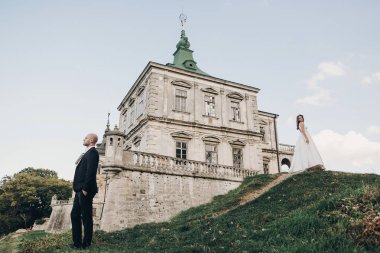 Parktaki eski şatonun yakınında güneş ışığında poz veren muhteşem gelin ve damat. Ayakta duran ve farklı taraflara bakan şık bir düğün çifti. Romantik an