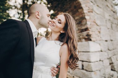 Parktaki eski şatonun yakınında akşam güneşinde kucaklaşan muhteşem gelin ve damat. Romantik bir an. Şık bir düğün çifti nazikçe sarılıp öpüşüyor.