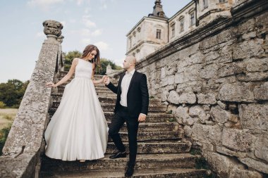 Muhteşem bir düğün çifti parktaki eski şatonun yakınındaki taş merdivenlerde yürüyor. Eski binanın arka planında poz veren muhteşem gelinlik ve damatla şık bir gelin.