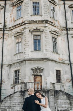 Güzel parktaki eski şatonun yakınında güneş ışığında kucaklaşan muhteşem bir düğün çifti. Tarz sahibi güzel gelin ve damat, antik yapı ve doğanın arka planında nazikçe sarılıyorlar.
