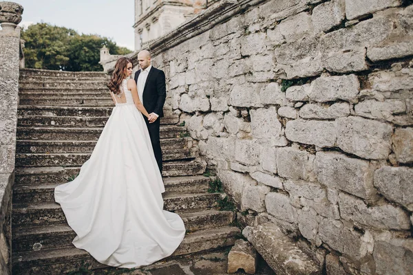 公園の古い城の近くの石の階段を歩く豪華な結婚式のカップル 驚くべきガウンと新郎のスタイリッシュな美しい花嫁は 古代の建物の背景にポーズ — ストック写真
