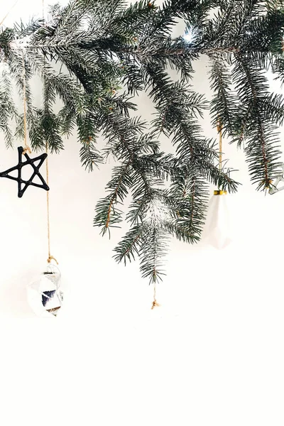 Stylish christmas ornaments hanging on fir branch on white wall.