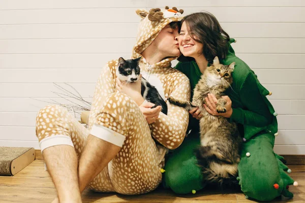 Happy couple in festive funny pajamas hugging their cats and smi — Stock Photo, Image