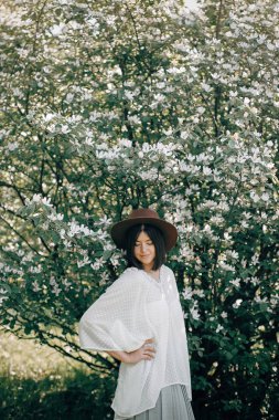 Beyaz flo ile çiçek açan ağaçta poz veren şapkalı şık boho kadın.