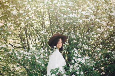 Beyaz flo ile çiçek açan ağaçta poz veren şapkalı şık boho kadın.