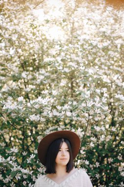 Beyaz flo ile çiçek açan ağaçta poz veren şapkalı şık boho kadın.