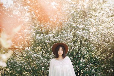 Beyaz flo ile çiçek açan ağaçta poz veren şapkalı şık boho kadın.
